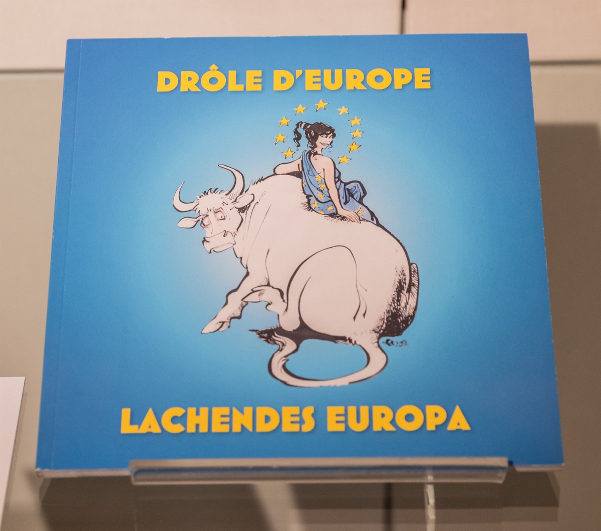 Eine Fotografie des Buches „Lachendes Europa“. Der Vordergrund zeigt die Europa, eine Figur der griechischen Mythologie, welche in einer blauen, mit Sternen verzierten Toga, grinsend auf dem weißen Zeus-Bullen sitzt. Ihr Kopf ist umgeben von den, in einem Kreis angeordneten, Sternen der EU-Flagge. Der Hintergrund ist hellblau, die Schrift gelb.
