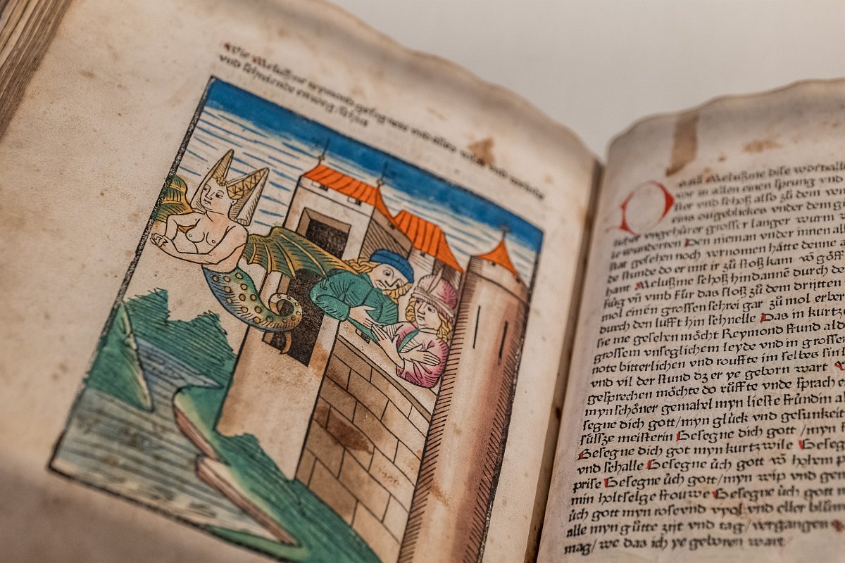 Zu sehen ist ein aufgeklapptes Buch, der Fokus liegt auf der Illustration auf der linken Seite. Diese zeigt zwei Ritter auf einer Burgmauer und eine Melusine, eine Wasserfee mit Schlangenleib, statt Beinen, und Drachenflügeln, welche gerade wegfliegt.