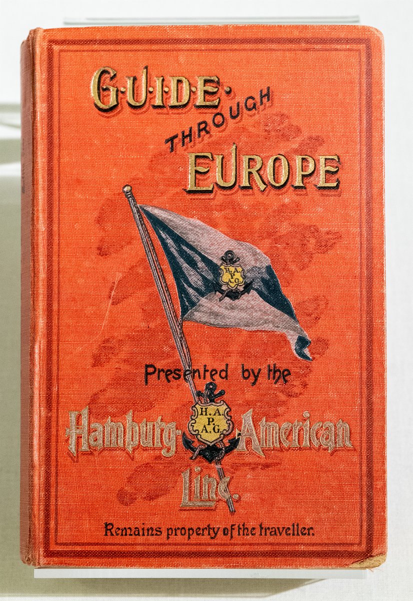 Buchumschlag „Guide through Europe“, roter Einband mit einer schwarzen Fahne, welche das Wappen der HAPAG (Hamburg-Amerikanische Packetfahrt-Actien-Gesellschaft) trägt.