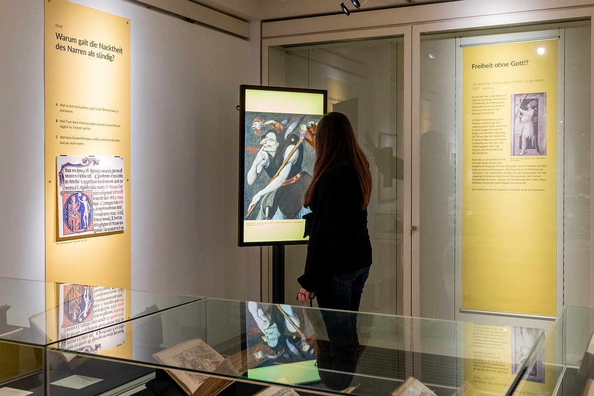 Auf dem Foto ist eine Ausstellungsbesucherin zu sehen, die den interaktiven Monitor bedient. Darauf abgebildet ist ein Narr mit verschiedenen typischen äußeren Merkmalen.