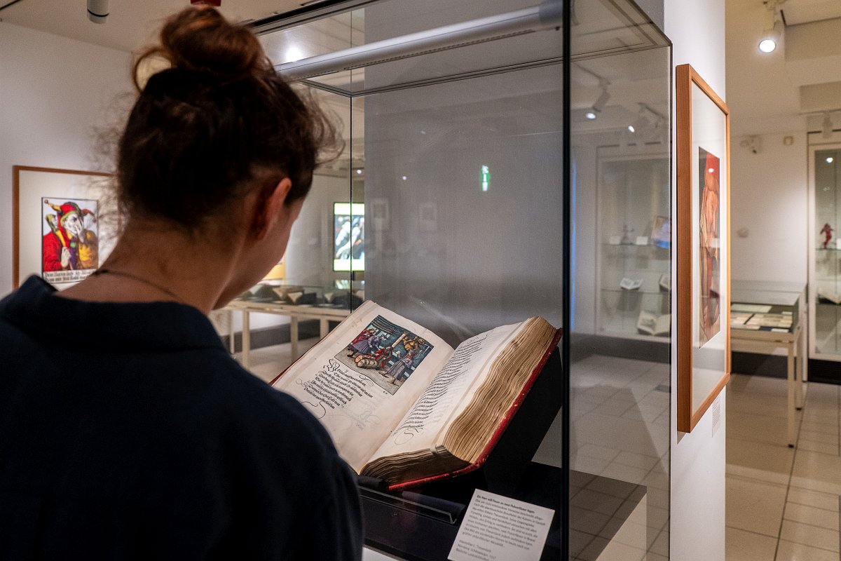 Das Foto zeigt eine Besucherin, die vor einer Glasvitrine steht. Darin liegt ein Buch mit der Graphik „Ein Narr will Feuer an zwei Pulverfässer legen“ (1517).