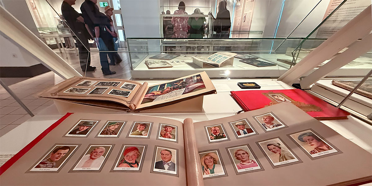 Aufnahme während der Kuratorenführung im Ausstellungsraum von „Wissen in Bildern – die bunte Welt der Sammelalben“. Im Vordergrund ist eine aufgeklappte Vitrine mit aufgeschlagenen Sammelalben der „Mercedes Filmbilder“-Reihe abgebildet. Im Hintergrund ist eine Gruppe von Menschen zu sehen, die um eine andere Vitrine stehen.