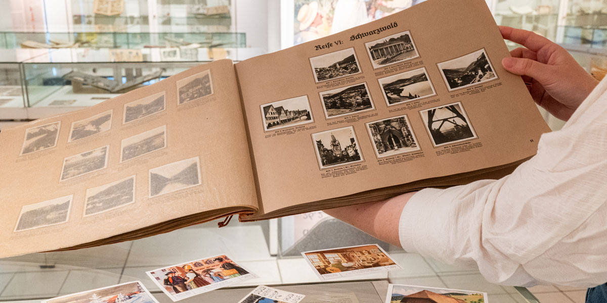 Eine Person hält im Ausstellungsraum eine Sammelalbum zum Thema „Schwarzwald“ in die Kamera. Darunter sind – auf einer Vitrine ausgebreitet– einzelne Sammelbilder zu erkennen.