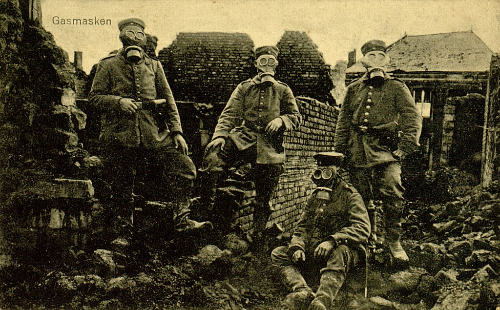 Zu sehen sind vier deutsche Soldaten im Feld. Sie tragen Gasmasken und stehen in einer zerstörten Siedlung. 