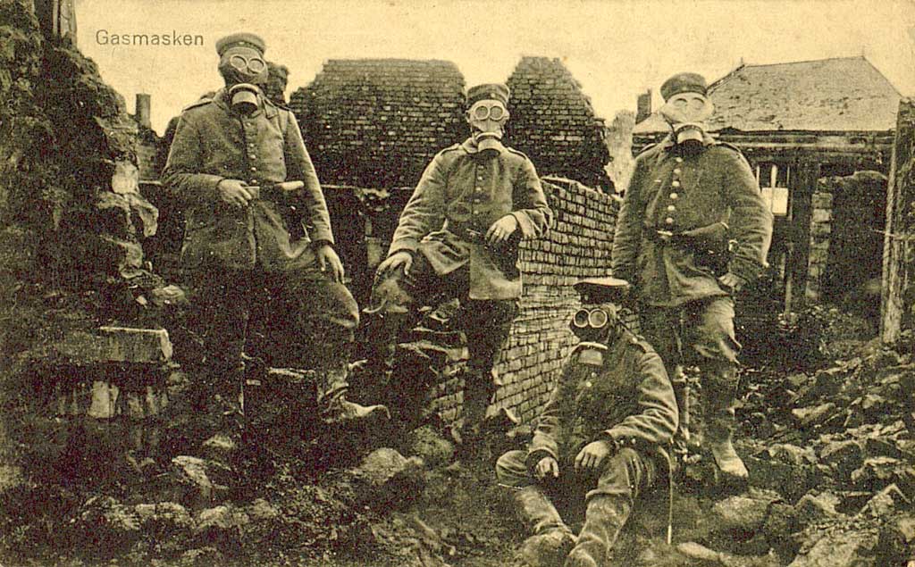 Zu sehen sind vier deutsche Soldaten im Feld. Sie tragen Gasmasken und stehen in einer zerstörten Siedlung. 