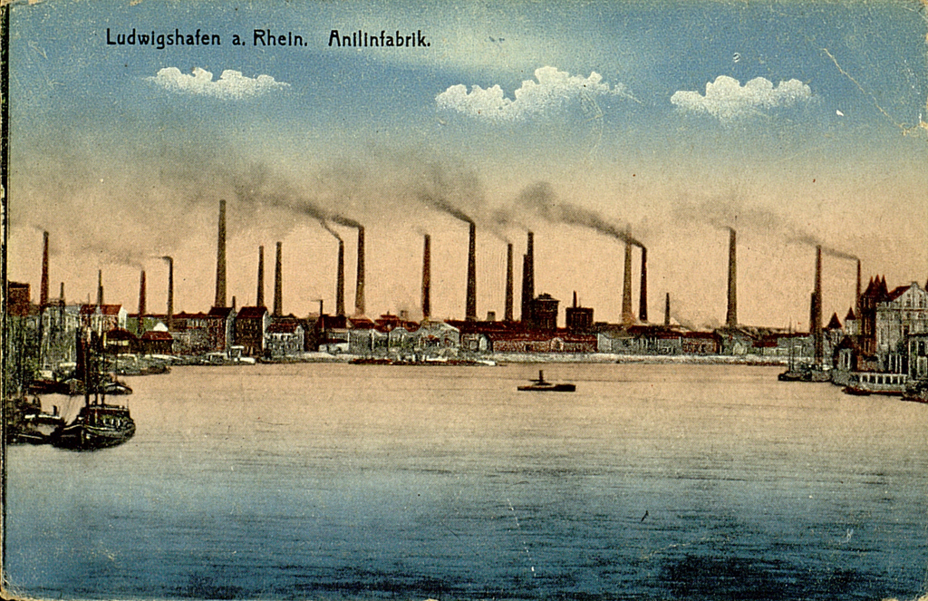 Zu sehen ist eine kolorierte Schwarz-Weiß Aufnahme der damaligen Anilinwerke in Ludwigshafen am Rhein. Die Fabrikanlage mit zahlreichenden rauchenden Schloten nimmt den Bildhintergrund ein. Im Vordergrund ist der Rhein zu sehen.  