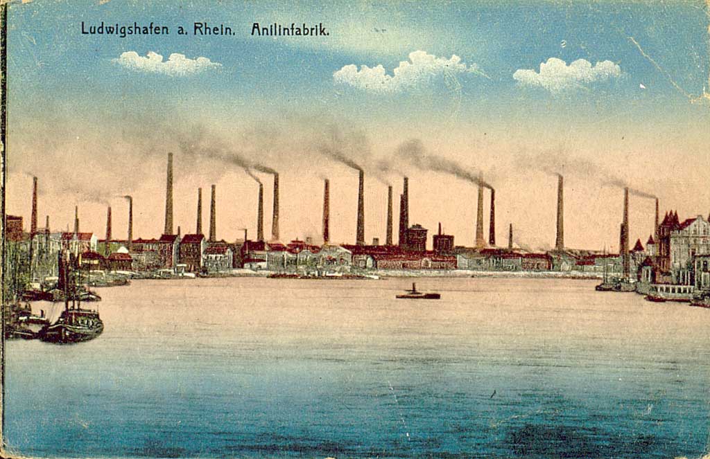 Zu sehen ist eine kolorierte Schwarz-Weiß Aufnahme der damaligen Anilinwerke in Ludwigshafen am Rhein. Die Fabrikanlage mit zahlreichenden rauchenden Schloten nimmt den Bildhintergrund ein. Im Vordergrund ist der Rhein zu sehen.  