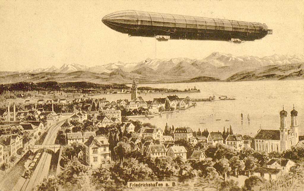 © Badische Landesbibliothek Karlsruhe Die Ansichtskarte zeigt einen Zeppelin über Friedrichshafen am Bodensee. Gut erkennbar ist im rechten Bildbereich die Schlosskirche, die Uferpromenade und die Kirche St. Nikolaus in der Mitte der Abbildung. Im Hintergrund, auf der anderen Seeseite sind die Alpen zu erkennen.