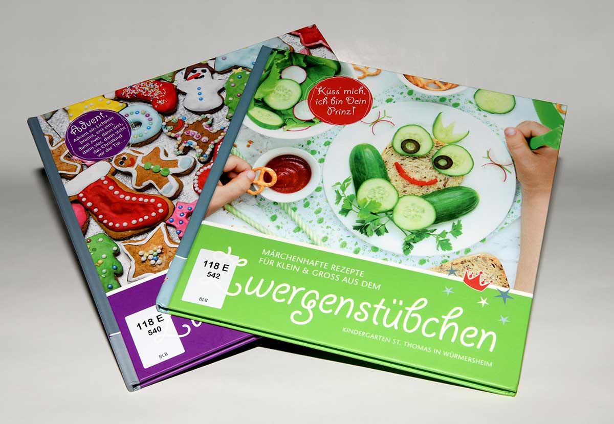 Zwei Kinderkochbücher mit bunten Titelseiten, eines zeigt einen Teller mit einem Frosch aus Gurkenscheiben und anderem Gemüse, das andere bunte verzierte Lebkuchen.