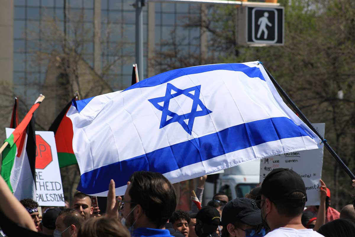 Das Foto zeigt eine Straßendemo mit palästinensischen Flaggen. Davor ragt eine eine israelische Flagge empor.