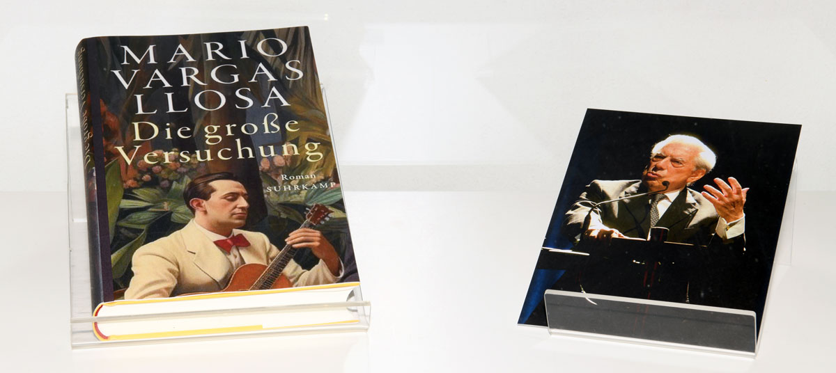Das Foto zeigt ein Buchcover des Romans „Die große Versuchung“ von Mario Vargas Llosa. Darauf ist ein junger Mann zu sehen, der Gitarre spielt. Er trägt einen weißen Smoking und eine rote Fliege. Rechts vom Buch wird ein Foto des Autors präsentiert. Er steht als älterer Mann am Rednerpult und gestikuliert.