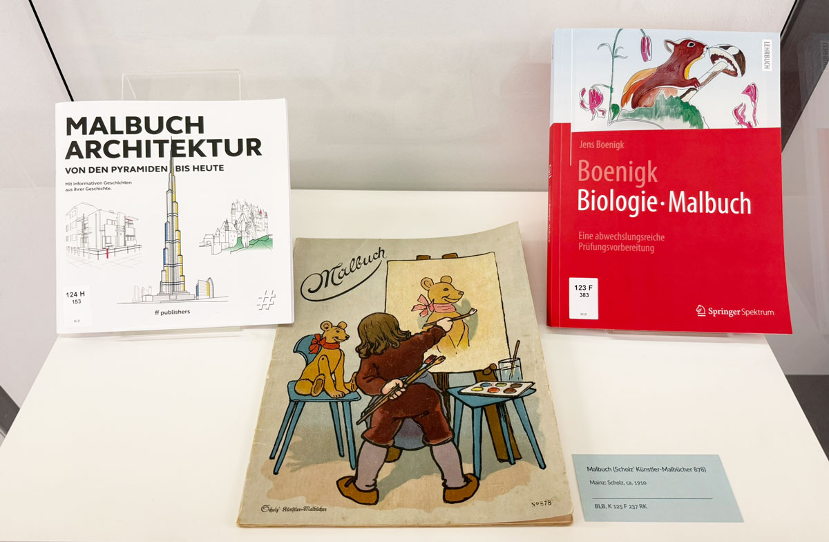 Auf dem Foto sind drei Malbücher abgebildet. Links „Malbuch Architektur“ mit Architekturskizzen auf dem Cover. In der Mitte „Scholzʹ Künstler-Malbücher 878“ mit einer Illustration, die ein Kind ausgestattet mit Farben und Pinseln vor einer Staffelei zeigt, daneben ein sitzender Teddy mit einer großen roten Schleife als Malmotiv. Rechts ist ein Biologie-Malbuch zu sehen, das Cover zeigt ein Eichhörnchen, das an einem Pilz riecht.