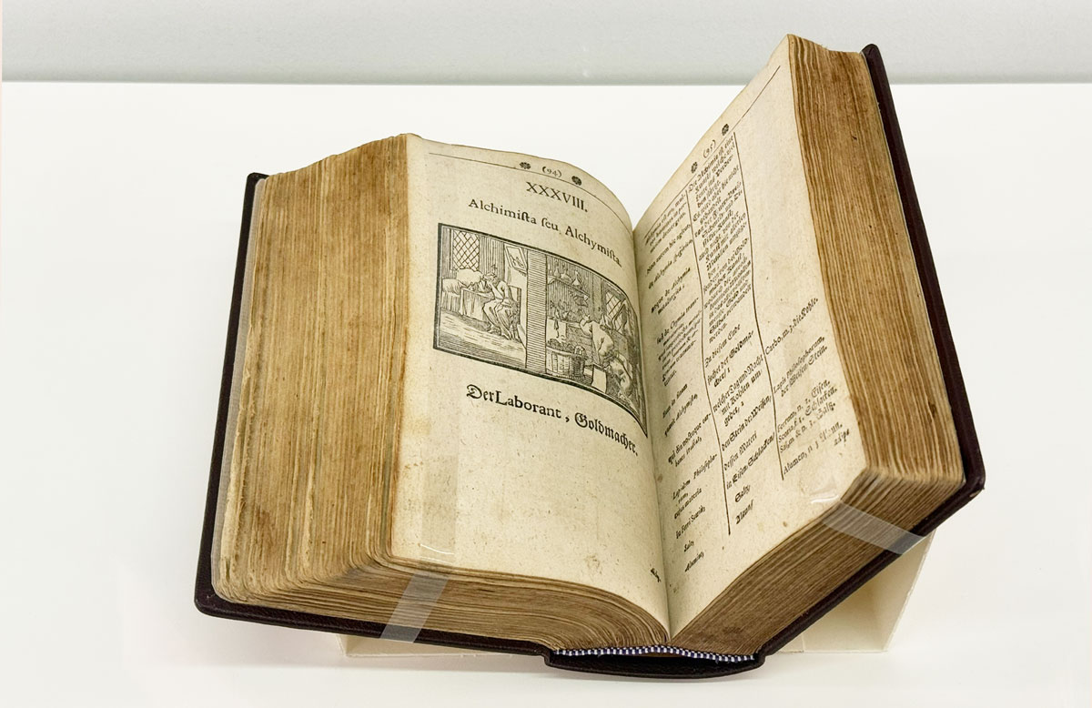 Zu sehen ist ein aufgeschlagenes Buch. Auf der linken Seite ist eine Illustration mit dem Untertitel „Der Laborant, Goldmacher.“ abgebildet.