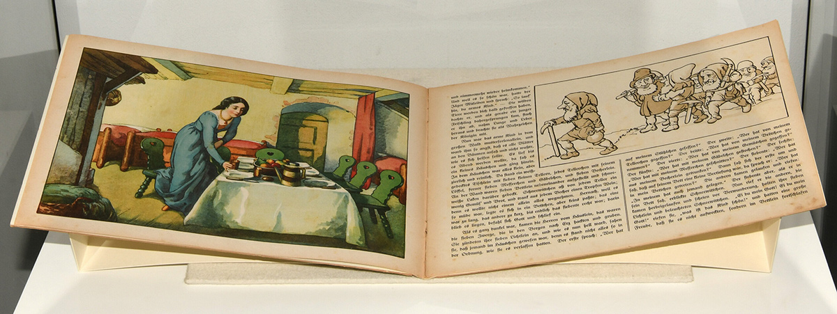 Zu sehen ist ein aufgeschlagenes Buch. Die linke Seite illustriert auf einer ganzseitigen Graphik, wie Schneewittchen den Tisch deckt. Auf der rechten Seite sind in einer kleineren Darstellung die sieben Zwerge zu sehen.