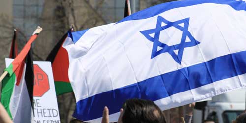 Das Foto zeigt eine Straßendemo mit palästinensischen Flaggen. Davor ragt eine eine israelische Flagge empor.