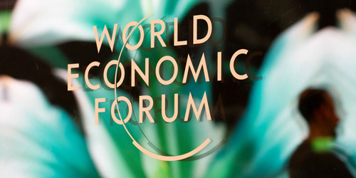 Zu sehen ist das Logo des „World Economic Forum“ auf einer Glasscheibe, im Hintergrund ist verschwommen der Kopf einer Person als Silhouette zu erkennen.