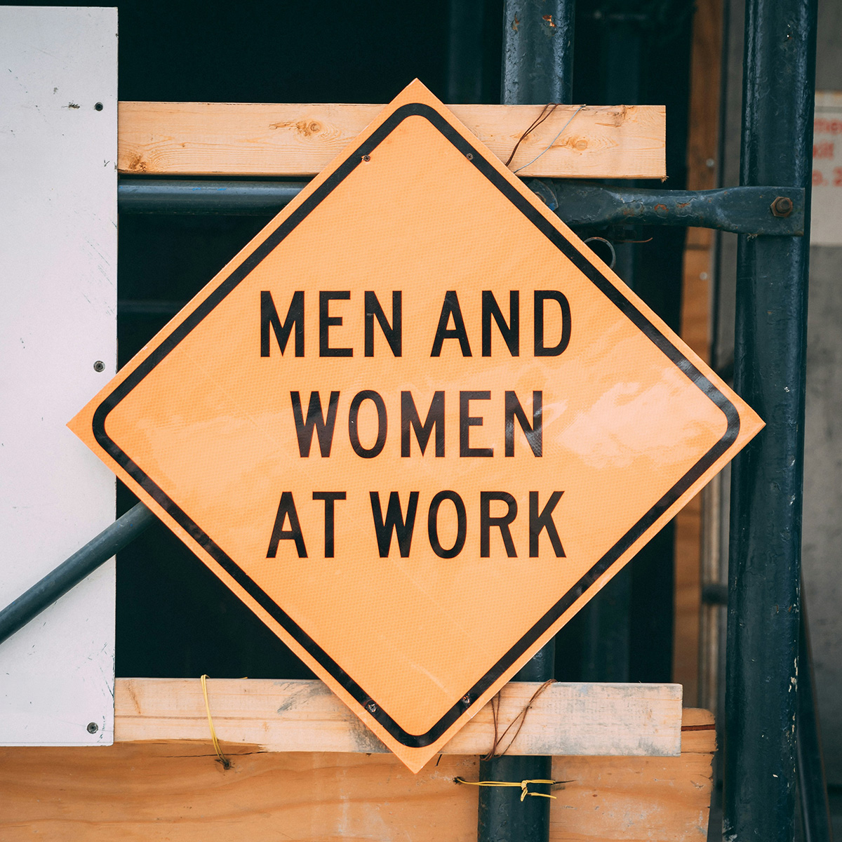 Zu sehen ist ein gelbes viereckiges Warnschild mit der Aufschrift „MEN AND WOMEN AT WORK“.