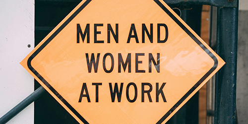 Zu sehen ist ein gelbes viereckiges Warnschild mit der Aufschrift „MEN AND WOMEN AT WORK“.