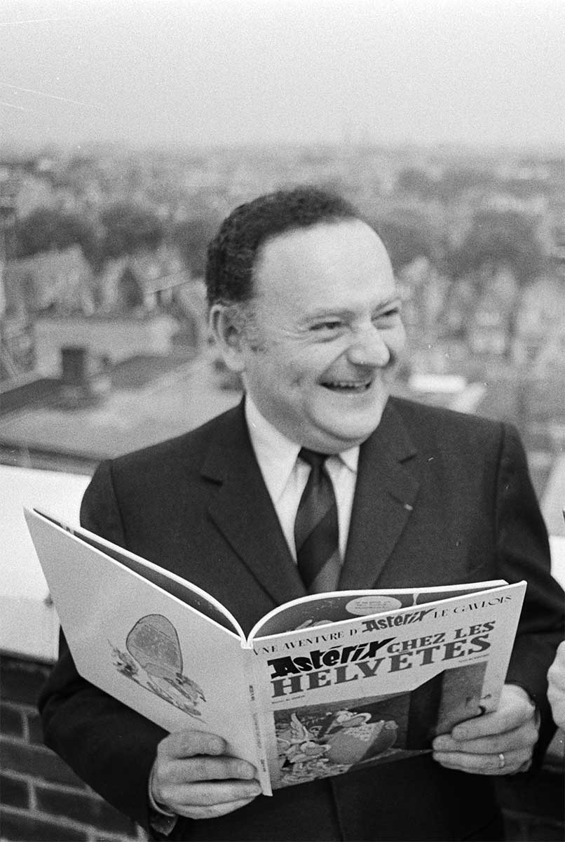 Goscinny steht im Anzug auf einer Dachterrasse und hält ein großformatiges Asterix-Comic mit dem Titel 'Astérix chez les Helvètes' in den Händen. Er lächelt und blickt leicht zur Seite. Im Hintergrund sind unscharf Dächer und Bäume einer Stadtlandschaft zu erkennen. Das Bild ist schwarz-weiß.