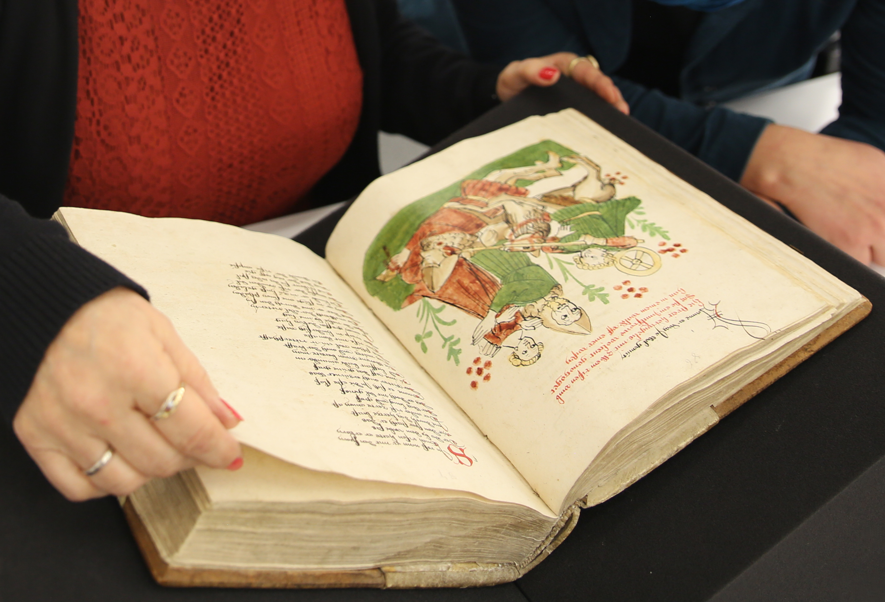 Das Bild zeigt zwei Personen, die sich die Wigalois-Handschrift ansehen. Aufgeschlagen sind die Blätter 43v und 44r mit einer Illustration, die Wigalois bei der Befreiung einer aus dem Artusreich geraubten Jungfrau zeigt.