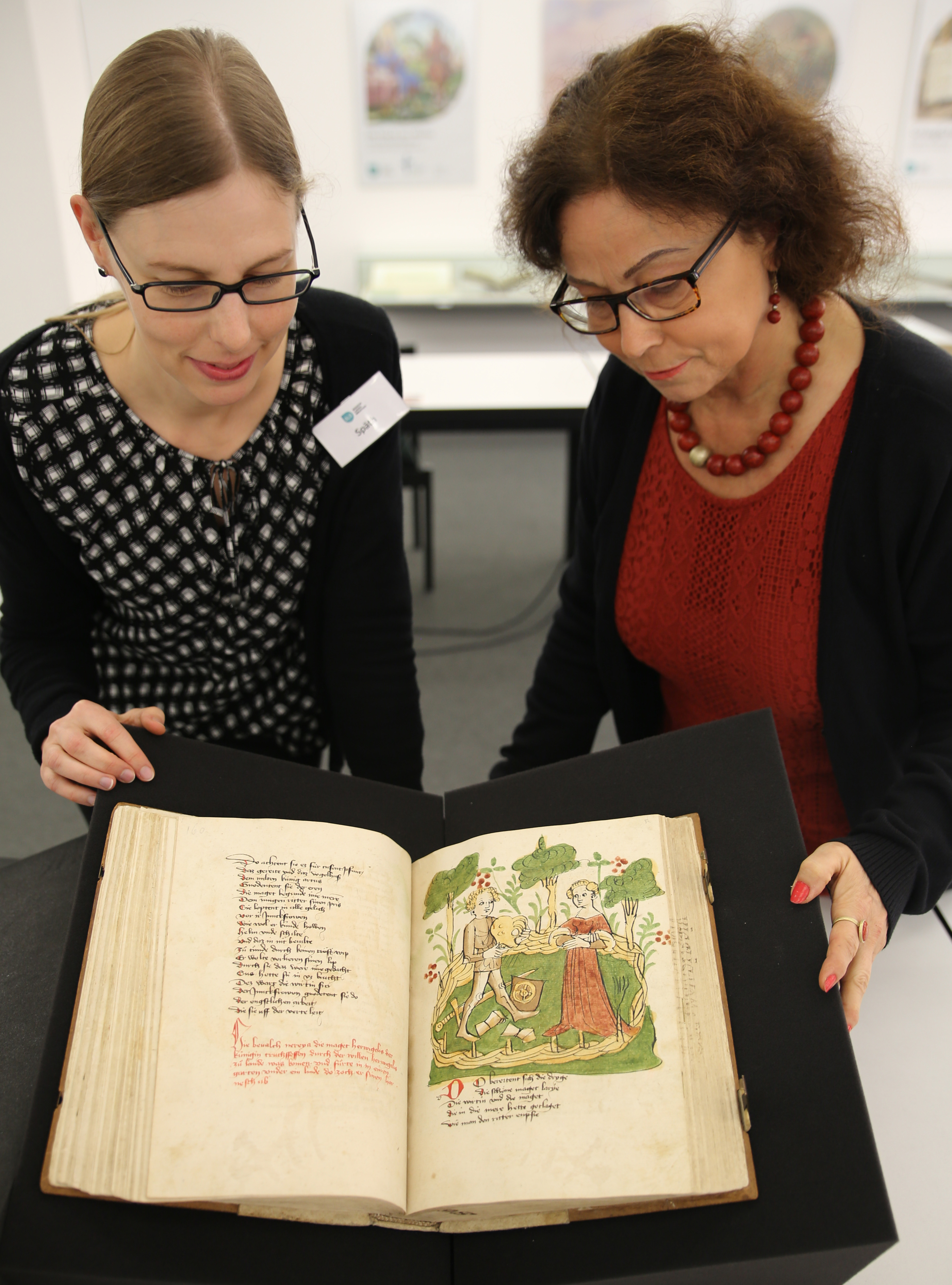 Zwei Frauen beugen sich über die aufgeschlagene Wigalois-Handschrift. Zu sehen ist die Illustration auf den Blättern 81v und 82r: Wigalois legt im Garten von Roimunt Rüstung und Waffen ab. 