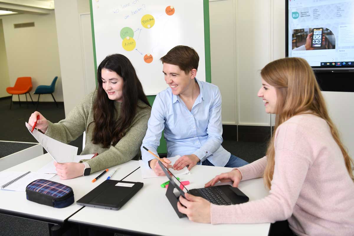 Zu sehen sind drei Personen in der Lernwerkstatt an einem Tisch mit unterschiedlichen Arbeitsmaterialien.