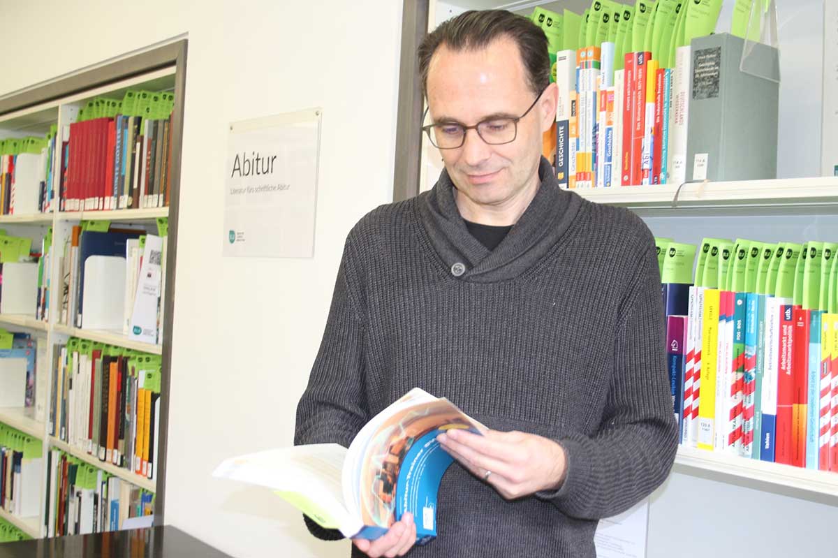 Auf dem Bild ist Ralf Weber vor dem physischen Abiturapparat, der ausgewählte Literatur zu den aktuellen Prüfungsthemen der allgemeinbildenden Gymnasien in Baden-Württemberg umfasst, zu sehen.