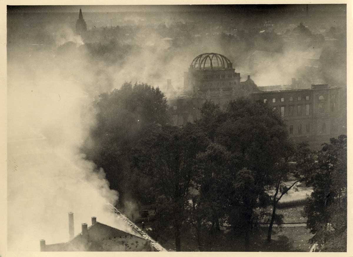 Die Fotografie aus der Bombennacht zeigt das brennende Sammlungsgebäude am Friedrichsplatz aus der Vogelperspektive, in Rauchschwaden gehüllt