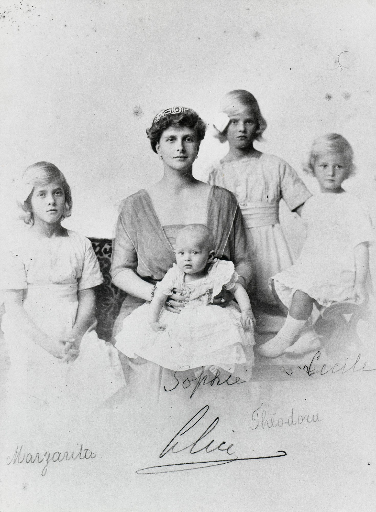 Zu sehen ist Prinzessin Alice von Griechenland mit ihren vier Töchtern. Prinzessin Alice sitzt auf einem Sofa mit dem Baby Sophia auf den Knieen. Prinzessin Margarita sitzt links neben ihr. Prinzessin Cecilia sitzt auf der Sofalehne rechts. Prinzessin Theodora steht zwischen ihrer Mutter und Prinzessin Cecilia.
