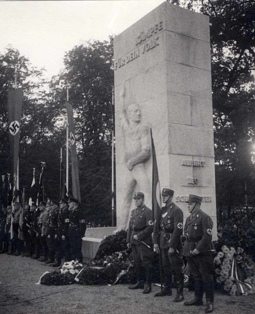 Zu sehen ist eine schwarz-weiße Fotografie der Einweihung des Schlageterdenkmals am 22. April 1934. Soldaten stehen in einer Reihe vor dem Denkmal, auf dem Boden, angelehnt an das Werk, sind große Blumenkränze zu finden.