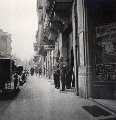 Der Gehweg einer Einkaufsstraße ist zu sehen. Links parken Autos, rechts daneben stehen Männer, teils in Uniform, vor Geschäftseingängen.