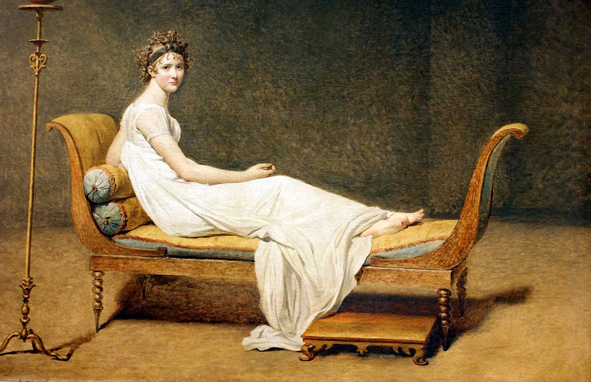 Eine junge Frau liegt halb aufgerichtet auf einer eleganten Liege. Sie trägt ein helles, schlichtes Kleid und blickt ruhig zur Seite. Der Hintergrund ist leer und betont ihre anmutige Haltung.
