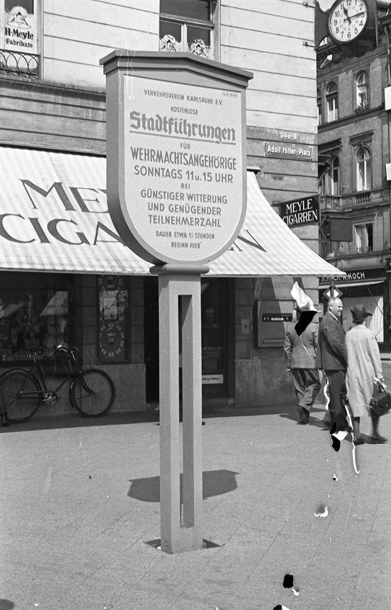 Straßenschild mit der Aufschrift 'Adolf Hitler Platz' an einer Gebäudeecke in einer belebten Fußgängerzone mit Passanten und Fahrrad.