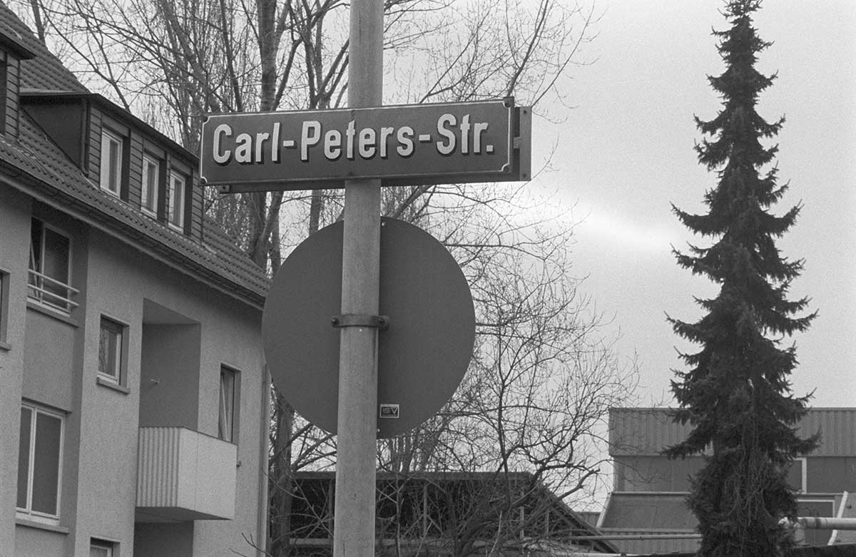 Straßenschild mit der Aufschrift 'Carl-Peters-Str.' an einem Mast vor einem Wohnhaus und kahlen Bäumen.