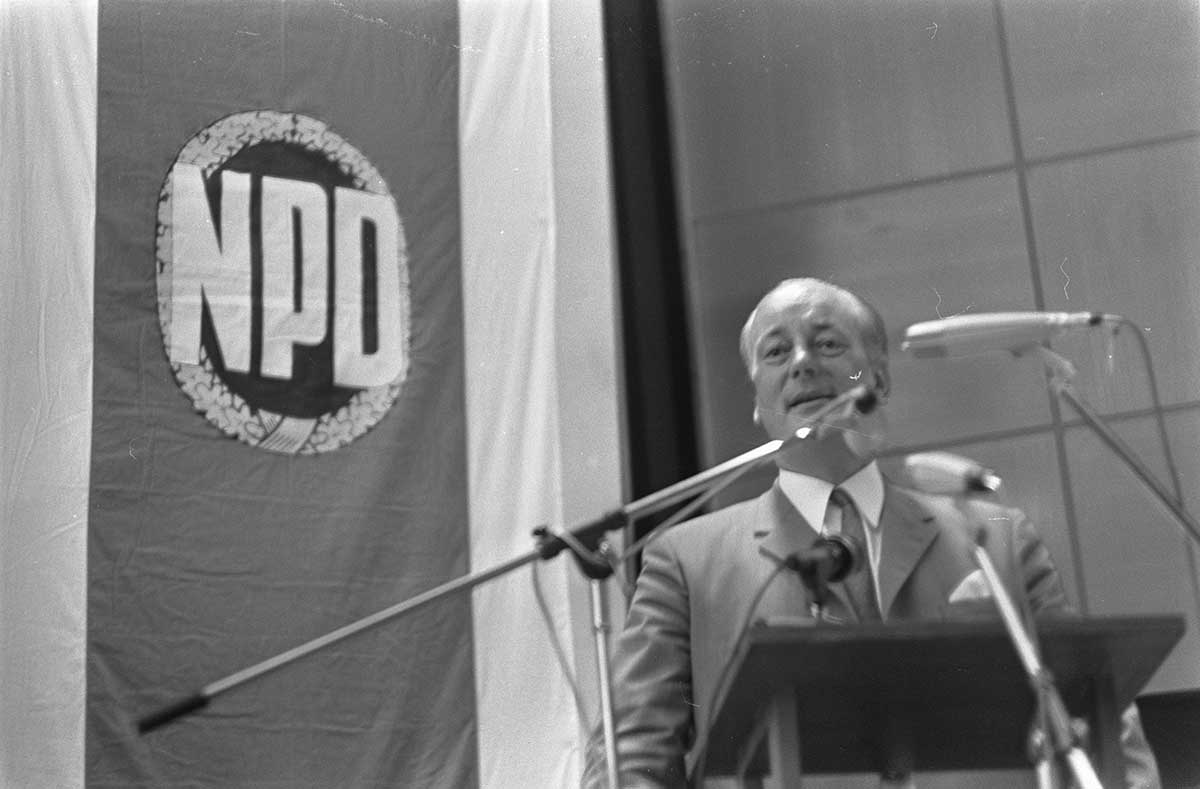 Adolf von Thadden spricht an einem Rednerpult vor einem Banner mit dem Schriftzug 'NPD'.