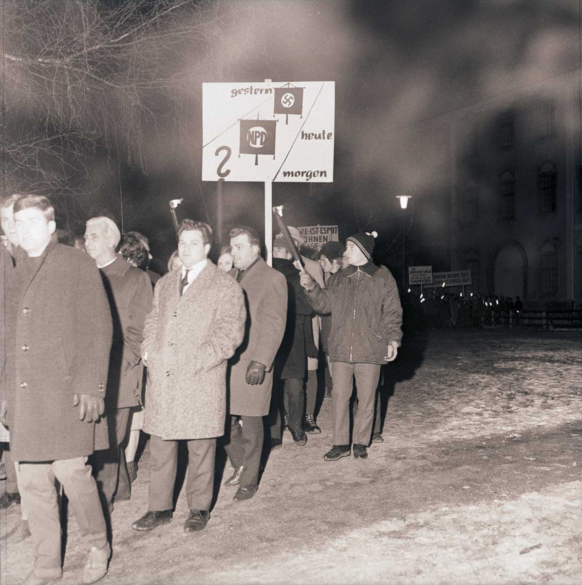 Gruppe von Menschen in Winterkleidung steht nachts im Freien, eine Person hält ein Schild mit den Worten 'gestern', 'heute', 'morgen' und Symbolen darunter.