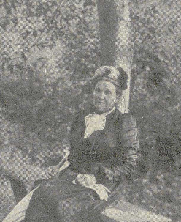Fotografie einer älteren Augusta Bender in dunklem Kleid mit weißem Kragen und Hut sitzt im Freien vor einem Baum.