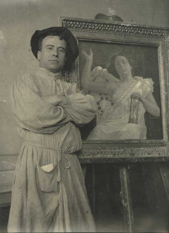Icilio Federico Joni steht mit verschränkten Armen vor einem Staffelei-Gemälde, trägt eine Malerjacke und einen Hut in seinem Atelier.