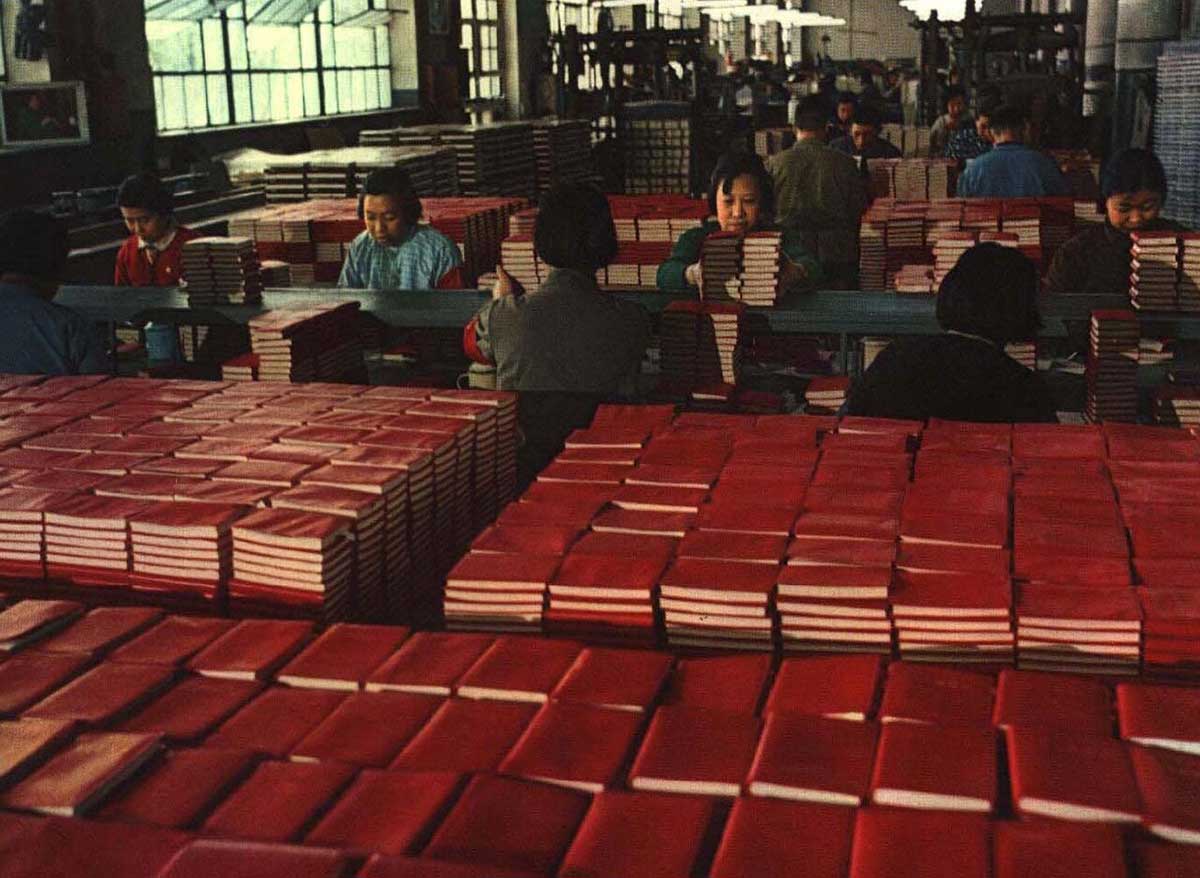 Blick in eine Druckerei mit mehreren Personen an Arbeitstischen und einem großen Stapel roter Bücher im Vordergrund.