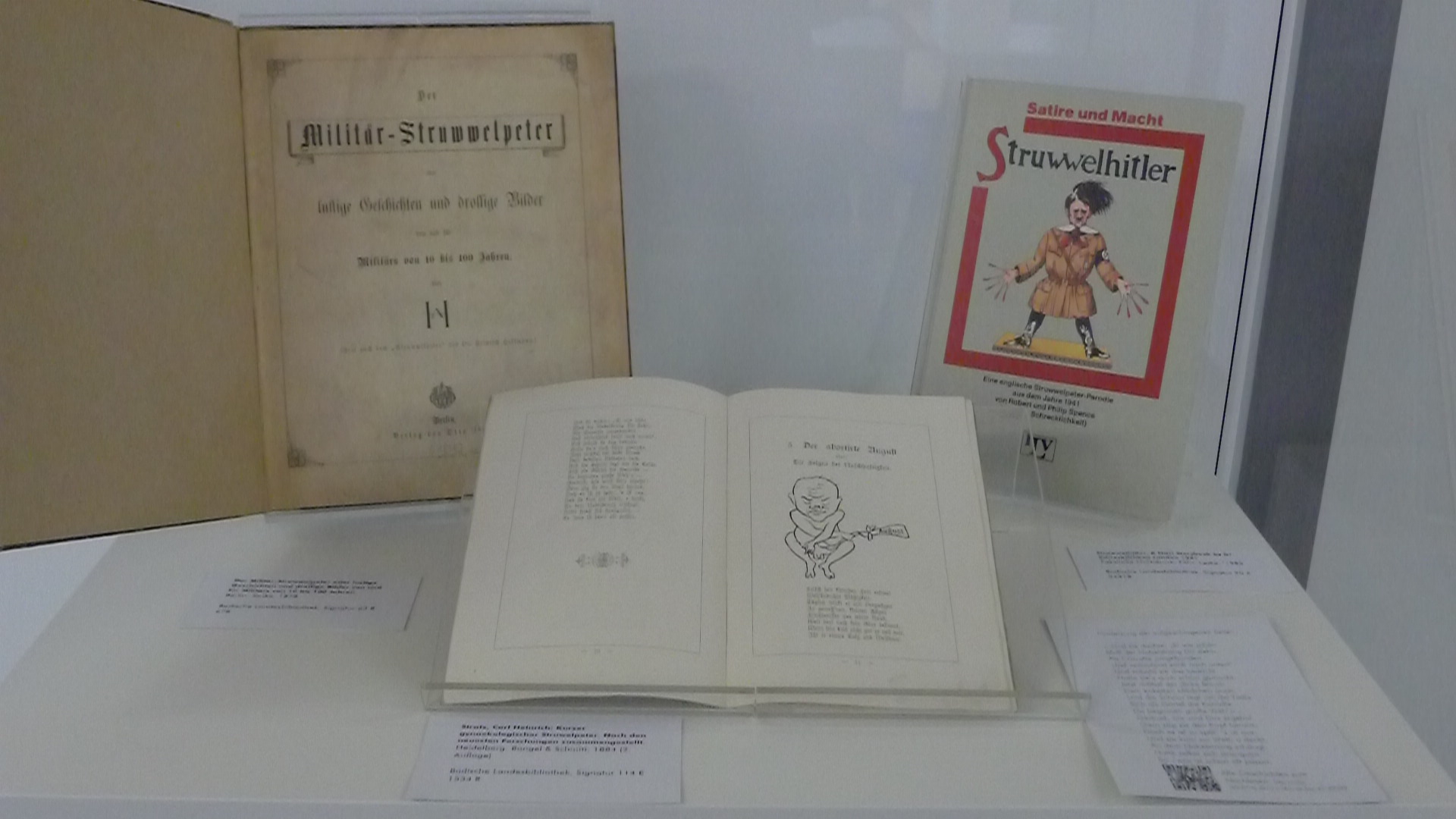 Ausgestellt in einer Vitrine sind drei Struwwelpetriaden. Links hinten das Deckblatt von "Militär-Struwwlpeter", rechts hinten das Cover von "Struwwlhitler" und vorne mittig eine Seite aus dem Buch "Kurzer gynaekologischer Struwelpeter" mit einer stilisierten Zeichnung eines "unartigen" Fötus.