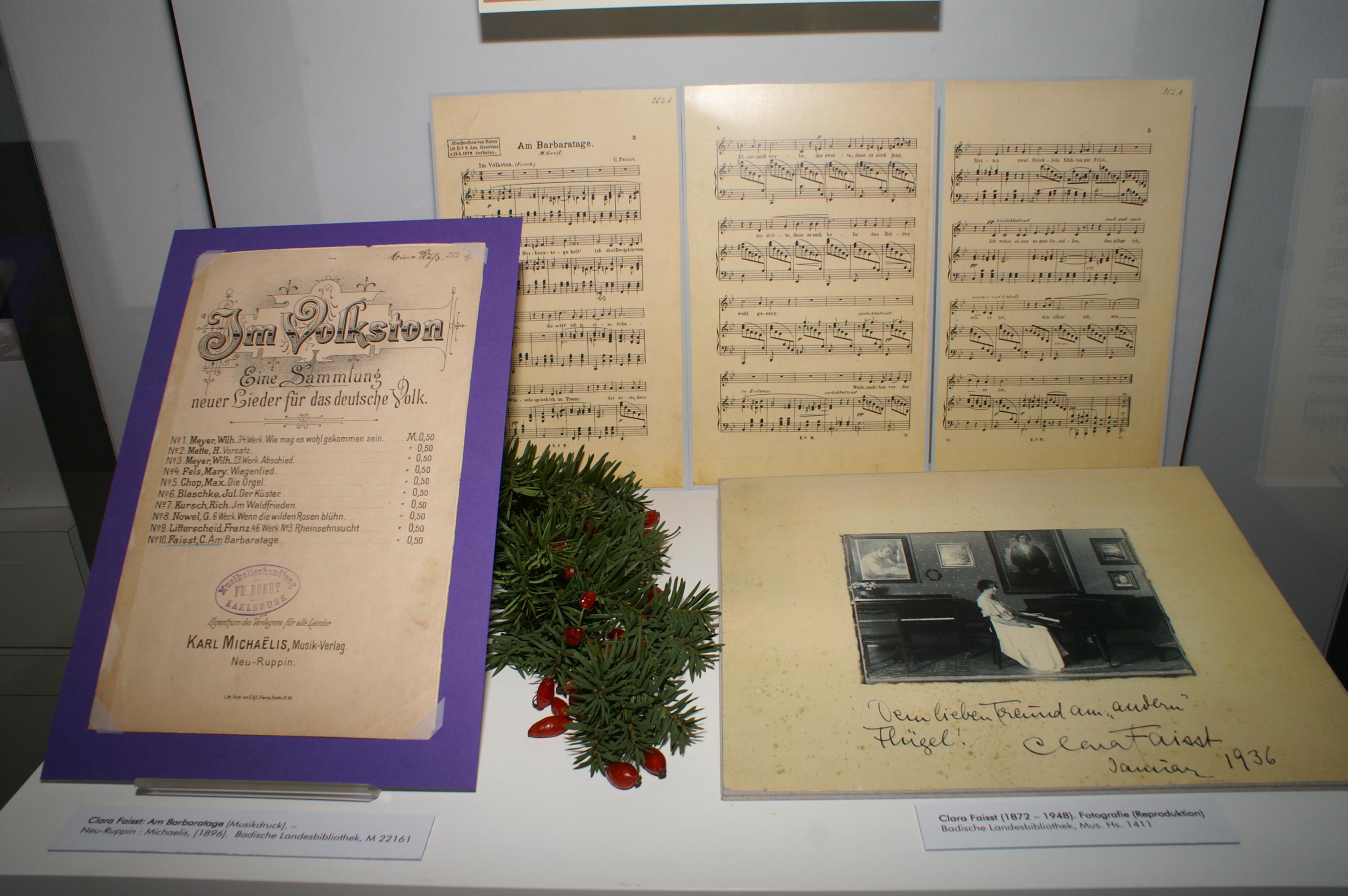 Ausgestellt sind Notenblätter, ein Musikdruck und eine schwarz-weiß Fotografie von Clara Faisst am Klavier. Dekoriert ist die Vitrine mit Tannenzweigen und Hagebutten.